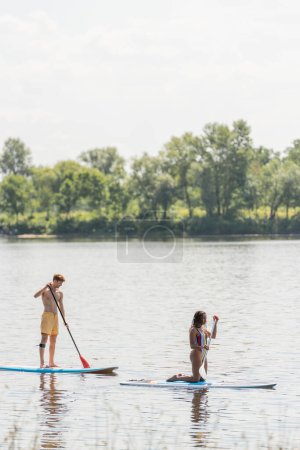 Photo for Side view of young redhead man and african american woman in colorful swimsuit spending summer weekend while sailing on sup boards on picturesque river - Royalty Free Image