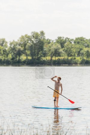 pełna długość sportowego i rudego człowieka w żółtych szortach pływackich stojących na desce z wiosłem i patrzących na jezioro z zielonymi drzewami na brzegu podczas letniego weekendu