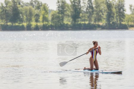 full length of cheerful and active african american woman in colorful swimsuit holding paddle and sailing on sup board along picturesque river bank in summer