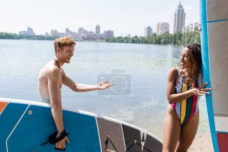 heureux rousse homme tenant conseil de repas et parler à enchanteresse afro-américaine en maillot de bain coloré près du lac et paysage urbain en arrière-plan en été