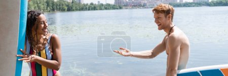 Foto de Hombre excitado y pelirrojo apuntando a la encantadora mujer afroamericana en traje de baño a rayas sosteniendo sup board y sonriendo cerca del lago en el día de fin de semana de verano, bandera - Imagen libre de derechos