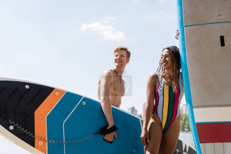 despreocupado pelirrojo hombre y deportivo africano americano mujer en colorido traje de baño celebración de sup boards, sonriendo y mirando hacia otro lado durante el fin de semana de verano cerca del río