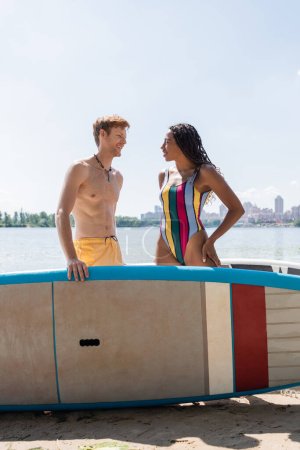 Photo for Smiling redhead man looking at brunette african american woman in colorful swimwear standing with hand on hip near sup board with river and cityscape on background - Royalty Free Image