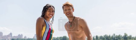 overjoyed and sportive redhead man and appealing african american woman in striped swimsuit looking at camera together under blue sky outdoors, banner