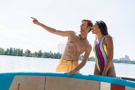 cheerful and redhead man looking away and pointing with finger near african american woman in colorful swimsuit while standing with sup boards near lake