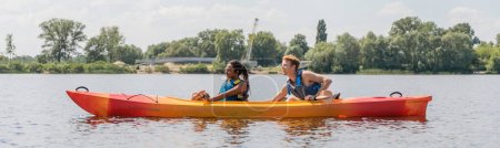 Photo for Side view of sportive and interracial couple in life vests sailing in kayak during summer water recreation on river with green picturesque bank, banner - Royalty Free Image