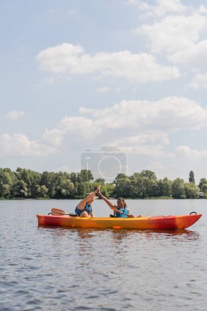 Foto de Despreocupados pareja interracial en chalecos salvavidas dando cinco altos durante el fin de semana de verano en el río y navegar en kayak deportivo a lo largo de la orilla verde del río bajo el cielo azul con nubes blancas - Imagen libre de derechos