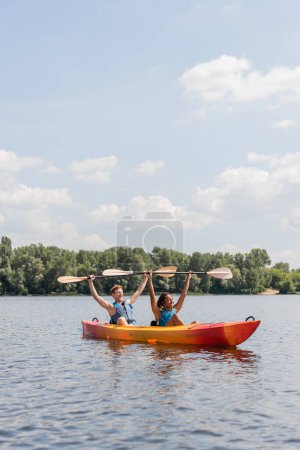 Ein paar fröhliche gemischtrassige Freunde in Schwimmwesten, die Paddel in erhobenen Händen halten, während sie im Kajak auf dem malerischen See unter blauem wolkenverhangenem Himmel mit grünem Flussufer im Hintergrund sitzen