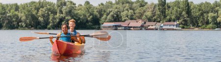 smiling redhead man and cheerful african american woman in life vests holding paddles while sailing in sportive kayak on lake with calm water and green picturesque shore, banner 