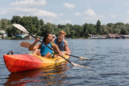 overjoyed african american woman and young redhead man in life vests spending summer weekend by sailing in kayak with paddles on lake with green scenery shore