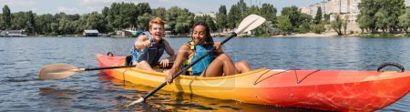 overjoyed redhead man pointing with finger near african american woman in life vest holding paddle while sailing in kayak along green riverside in summer, banner