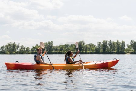 Photo for Side view of smiling redhead man and brunette african american woman in life vests spending summer weekend by sailing in kayak on river under cloudy sky in summer - Royalty Free Image