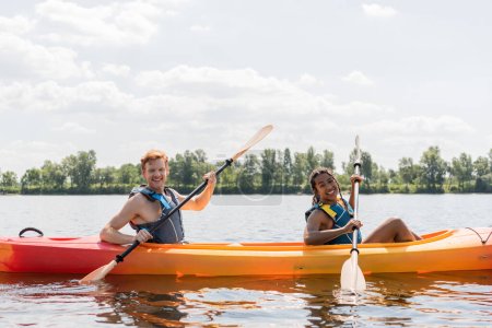 pretty african american woman and young redhead man in life vests smiling at camera while spending summer weekend on river and sailing in kayak with paddles under blue cloudy sky