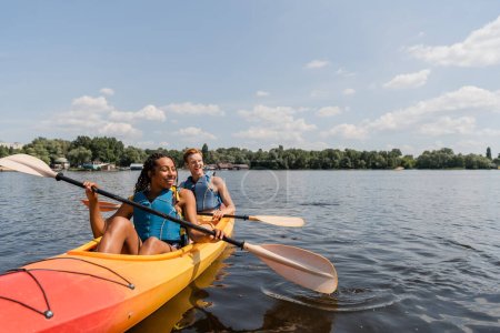Photo for Carefree and charming african american woman and young redhead man in life vests sailing in sportive kayak on calm water surface under blue sky with clouds on summer day - Royalty Free Image