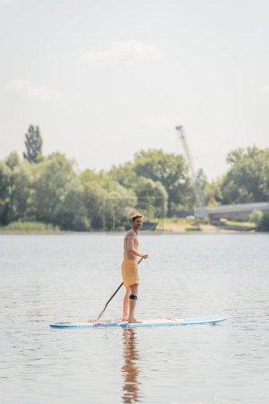 full length of sportive redhead man in yellow swim shorts holding paddle and looking at camera while sailing on picturesque lake on summer weekend day
