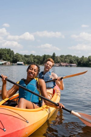 aufgeregtes multiethnisches Paar in Schwimmwesten verbringt Sommerwochenende auf malerischem See beim Paddeln im sportlichen Kajak unter blauem Himmel mit weißen Wolken