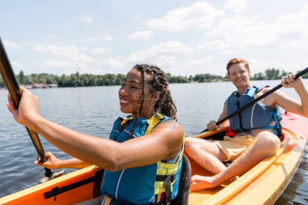 Photo for Cheerful african american woman in life vest sailing in kayak while spending time on river with young and redhead man paddleboarding on blurred background - Royalty Free Image