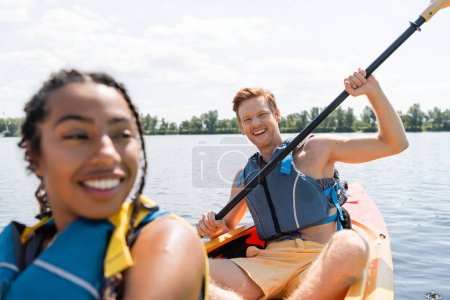 Foto de Hombre pelirrojo lleno de alegría en chaleco salvavidas sentado en kayak y sosteniendo la paleta mientras mira la cámara cerca de la mujer afroamericana sonriendo en primer plano borroso - Imagen libre de derechos