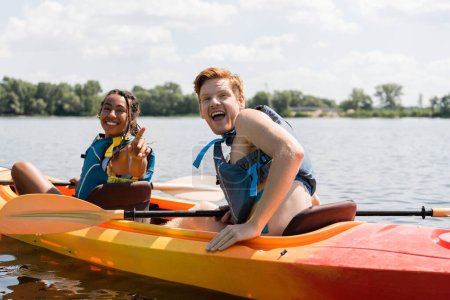 hombre pelirrojo excitado con la boca abierta mirando a la cámara cerca de la alegre mujer afroamericana en chaleco salvavidas señalando con el dedo mientras navega en kayak en el lago en verano