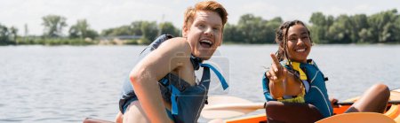 joven y excitado pelirrojo con la boca abierta mirando a la cámara cerca de la mujer afroamericana llena de alegría en chaleco salvavidas señalando con el dedo mientras navega en kayak en el día de verano, pancarta