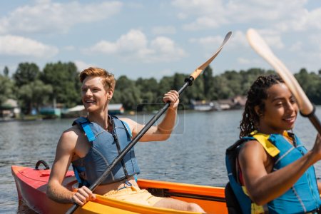 Téléchargez les photos : Excité rousse homme dans la vie gilet regardant loin tout en naviguant en kayak sportif avec jolie femme afro-américaine pendant les loisirs loisirs sur le lac en été - en image libre de droit