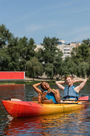 Foto de Complacida y descalza mujer afroamericana y pelirroja sonriente en chalecos salvavidas relajándose con los ojos cerrados en kayak deportivo durante el fin de semana de verano en el lago - Imagen libre de derechos