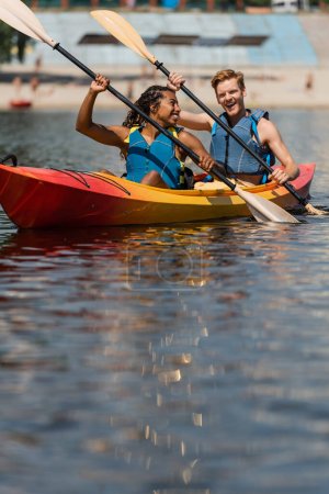 encantadora mujer afroamericana en chaleco salvavidas mirando al hombre pelirrojo excitado mientras navega en kayak deportivo con paletas en el día de verano en primer plano borrosa