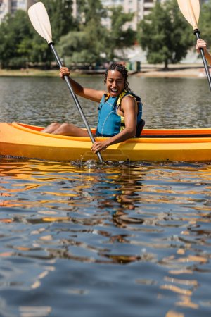 Photo for Impressed and cheerful african american woman in life vest holding paddle while sailing in sportive kayak during recreation weekend on city lake on summer day - Royalty Free Image