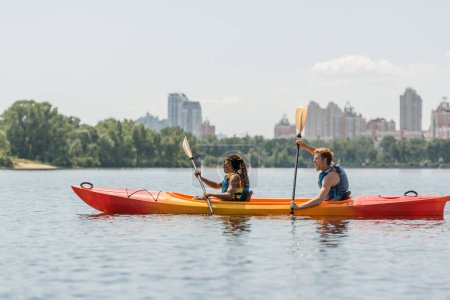 Photo for Side view of active african american woman and excited redhead man in life vests paddling in sportive kayak along riverside with picturesque cityscape on blurred background - Royalty Free Image