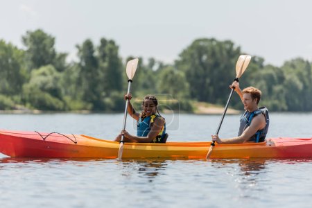 joyful and active interracial couple in life vests paddling in sportive kayak while spending time on lake with blurred green shore during summer vacation