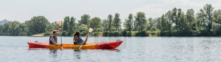 Photo for Side view of young man and african american woman in life vests sailing in sportive kayak during active summer weekend on picturesque river with green trees on bank, banner - Royalty Free Image