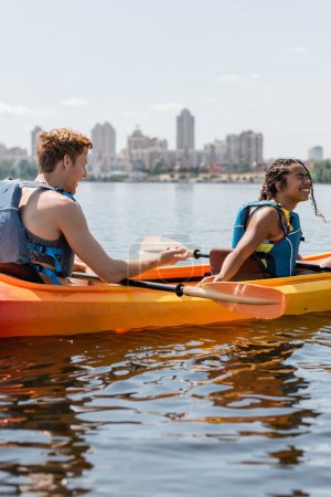 jeune homme rousse dans la vie gilet parlant à la femme afro-américaine ravie rire les yeux fermés et assis en kayak sportif sur la rivière avec paysage urbain flou sur le fond