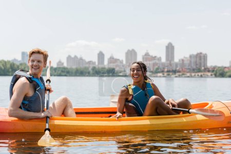 african american woman and redhead man in life vests sitting in sportive kayak with paddles and looking away on lake with picturesque cityscape on blurred background