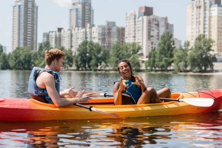 cheerful african american woman in life vest gesturing near young redhead man and having fun in sportive kayak on lake with modern buildings on background