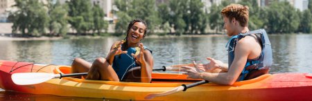 fröhliche afrikanisch-amerikanische Frau in Schwimmweste gestikulierend in der Nähe eines jungen rothaarigen Mannes, während sie das Sommerwochenende auf dem Fluss verbringt und im sportlichen Kajak sitzt, Banner