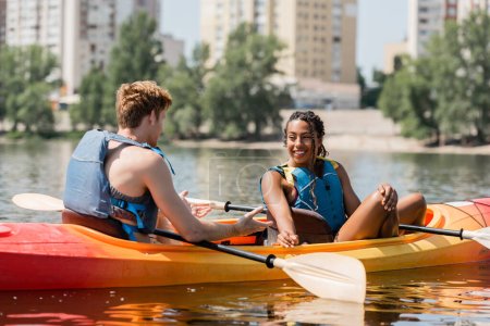 appealing african american woman looking at young redhead man in life vest and talking in sportive kayak on summer weekend with city scape on blurred background