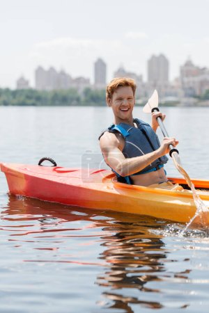 young and overjoyed redhead man in life vest looking at camera and holding paddle while sailing in sportive kayak on lake with cityscape on blurred background