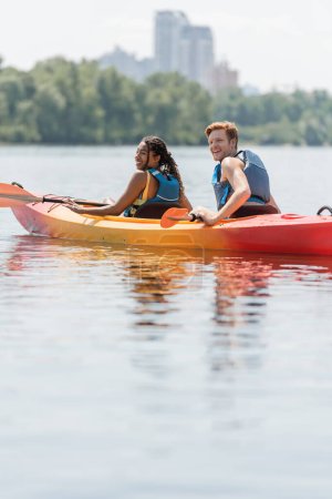 happy african american woman and young, smiling redhead man in life vests sitting in kayak near paddles and looking away during summer weekend on calm river