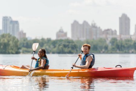 encantadora mujer afroamericana y el hombre pelirrojo deportivo en chalecos salvavidas mirando a la cámara durante la navegación en kayak a lo largo de la orilla del río con paisaje urbano borroso