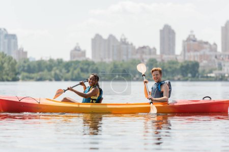 Photo for Positive and sportive interracial couple in life vests sitting with paddles in kayak and smiling at camera on river with picturesque cityscape on blurred background - Royalty Free Image