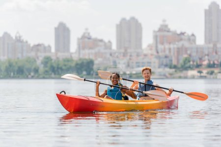 happy and impressed multiethnic friends in life vests holding paddles and smiling at camera in sportive kayak on river with scenic cityscape on blurred background