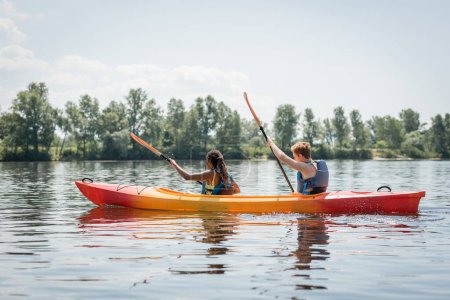 Foto de Vista lateral de pareja multiétnica activa en chalecos salvavidas sosteniendo paletas mientras navega en kayak deportivo en el río con pintoresca orilla con árboles verdes en verano - Imagen libre de derechos