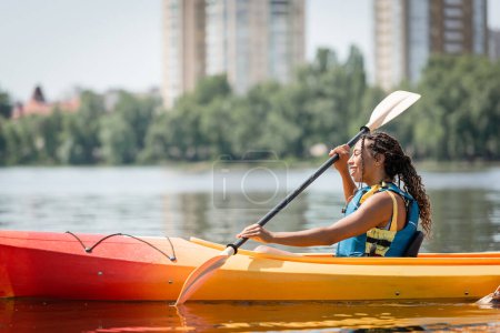 Photo for Side view of active and charming african american woman in life vest sailing in sportive kayak with paddle on summer weekend day on blurred background - Royalty Free Image