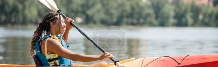 Photo for Side view of active and happy african american woman in life vest holding paddle and sailing in kayak while spending summer weekend on scenic river, banner - Royalty Free Image