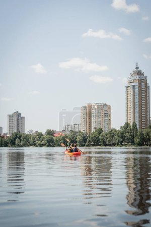 back view of interracial sportive couple in life vests sailing in kayak along riverside with green trees and contemporary buildings during recreation activity on summer weekend  