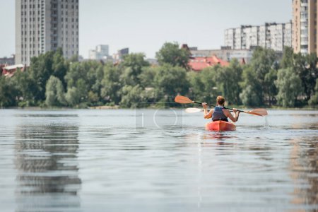 vista trasera de la pareja interracial deportiva en chalecos salvavidas que navegan en kayak cerca de la orilla del río con árboles verdes y edificios modernos de la ciudad durante el fin de semana de verano