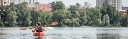 back view of active multiethnic friends in life vests sailing in sportive kayak with paddles on city river with green bank during recreation weekend in summer, banner