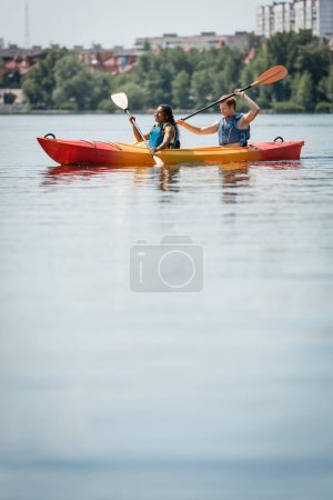 mujer afroamericana feliz y joven hombre activo en chalecos salvavidas pasar tiempo en el lago de la ciudad y navegar en kayak deportivo con paletas en primer plano borrosa