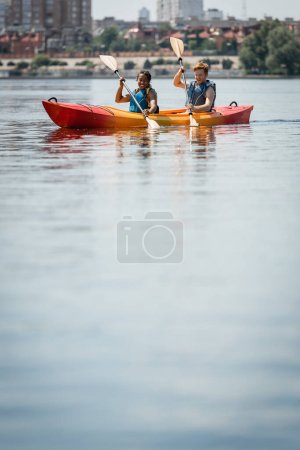 positive and active multiracial couple in life vests holding paddles while sailing in sportive kayak on calm water surface during summer recreation weekend on city lake