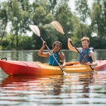 Charming african american woman with young and redhead man in life vests smiling while paddling in sportive kayak on lake with green trees on shore in summer